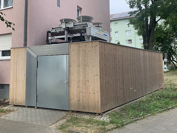Foto, Wärmepumpenanlage mit zwei Ventilatoren auf einem Holzschuppen neben einem rosa Wohngebäude