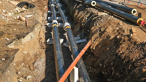 Foto, Zwei parallele schwarze Rohre mit weißen Verbindungsstücken liegen in einem Erdgraben, daneben ein orangefarbenes Rohr, umgeben von Erde und Kies.