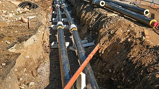 Foto, Zwei parallele schwarze Rohre mit weißen Verbindungsstücken liegen in einem Erdgraben, daneben ein orangefarbenes Rohr, umgeben von Erde und Kies.