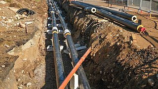 Foto, Zwei parallele schwarze Rohre mit weißen Verbindungsstücken liegen in einem Erdgraben, daneben ein orangefarbenes Rohr, umgeben von Erde und Kies.