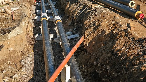 Foto, Zwei parallele schwarze Rohre mit weißen Verbindungsstücken liegen in einem Erdgraben, daneben ein orangefarbenes Rohr, umgeben von Erde und Kies.