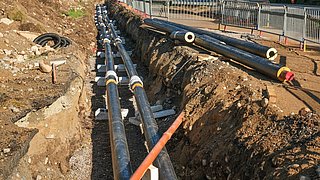 Foto, Zwei parallele schwarze Rohre mit weißen Verbindungsstücken liegen in einem Erdgraben, daneben ein orangefarbenes Rohr, umgeben von Erde und Kies.