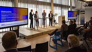 Foto, eine Podiumsdiskussion findet in einem Raum mit vier Rednern an Stehtischen und einem Publikum auf Stühlen statt.