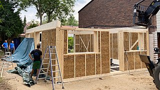 Foto, Rohbau eines Hauses aus Stroh und Holz mit drei Arbeitern und Baugeräten im Garten neben einem bestehenden Haus