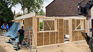 Foto, Rohbau eines Hauses aus Stroh und Holz mit drei Arbeitern und Baugeräten im Garten neben einem bestehenden Haus