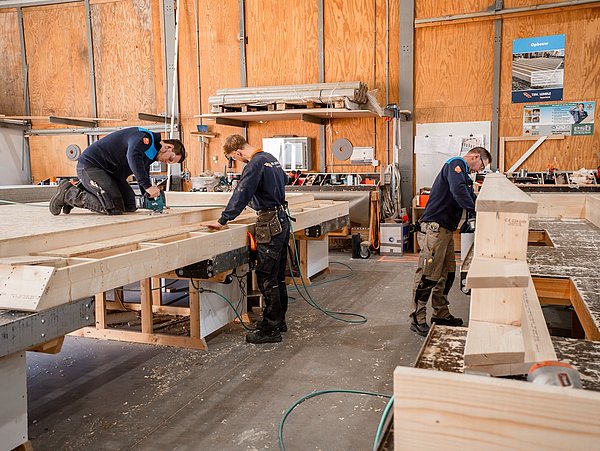 Foto, drei Handwerker bearbeiten Holzbalken in einer Werkhalle mit Werkzeugen und Arbeitskleidung.