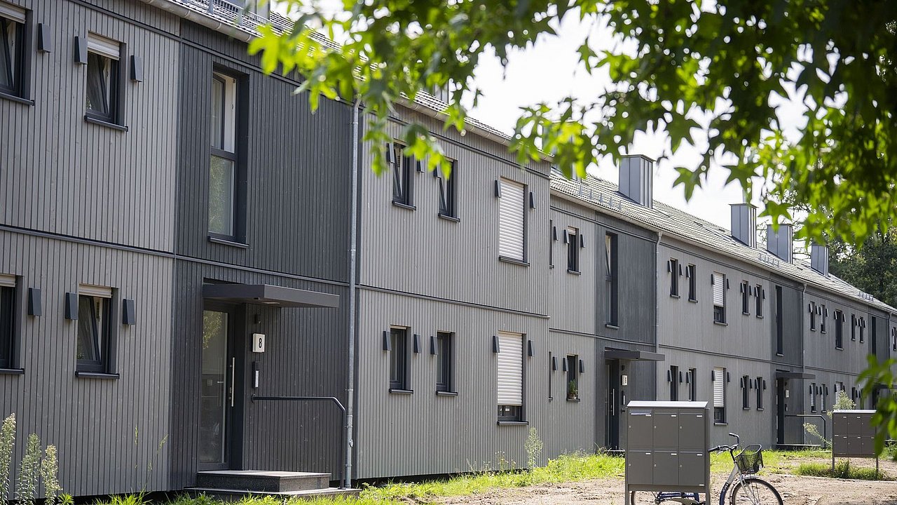 Foto, Reihe moderner, grauer Reihenhäuser mit vertikaler Holzverkleidung und mehreren Fenstern, davor Briefkästen und ein Fahrrad