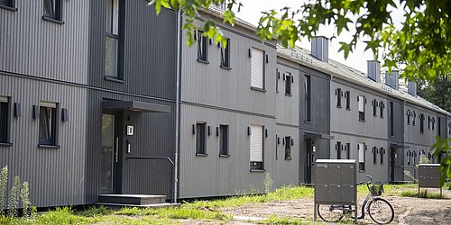 Foto, Reihe moderner, grauer Reihenhäuser mit vertikaler Holzverkleidung und mehreren Fenstern, davor Briefkästen und ein Fahrrad