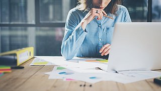 Foto, Blick auf einen Schreibtisch, im Vordergrund Aktenordner, Dokumenten und ein aufgeklappter Laptop. Hinter dem Laptop Oberkörper einer Person, die am Schreibtisch sitzt.