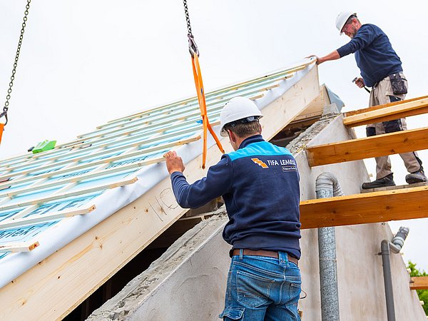 Foto, zwei Bauarbeiter mit Schutzhelmen montieren ein Dach mit Holzlatten und Dämmmaterial.