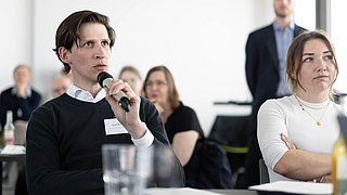 Foto, Mann mit Mikrofon und Namensschild sitzt an einem Tisch mit Glas und Unterlagen in einem hellen Raum mit mehreren Personen im Hintergrund.