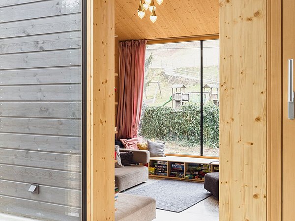 Foto, Übergang von einem Raum mit Holzvertäfelung zu einem Wohnzimmer mit großen Fenstern, grauem Sofa und Bücherregal