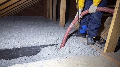 Foto, Person in Arbeitskleidung füllt mit Schlauch Dämmmaterial auf einem Dachboden auf Holzbalken ab.