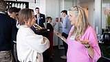 Foto, Zwei Frauen im Gespräch, eine trägt ein helles Oberteil und die andere ein pinkfarbenes Oberteil, im Hintergrund mehrere Personen in einem modernen Innenraum mit Glaswänden.