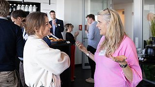 Foto, Zwei Frauen im Gespräch, eine trägt ein helles Oberteil und die andere ein pinkfarbenes Oberteil, im Hintergrund mehrere Personen in einem modernen Innenraum mit Glaswänden.