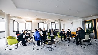 Foto, Mehrere Personen sitzen in einem modernen Büro auf bunten Sesseln und Stühlen in einem Kreis und unterhalten sich.