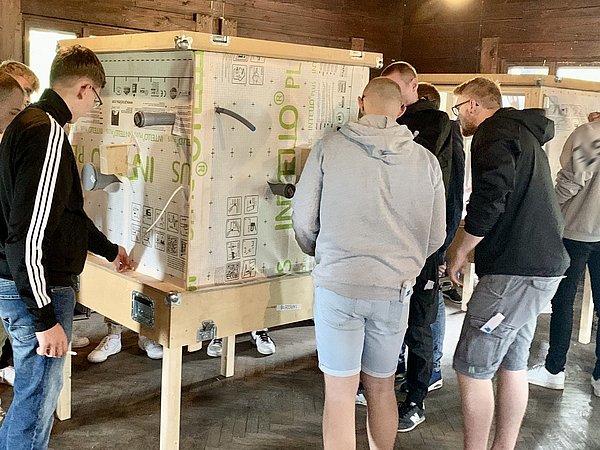Gruppe von Handwerker-Azubis, die an mehreren Holzrahmen mit Plänen und Zeichnungen arbeiten. Sie tragen Freizeitkleidung.