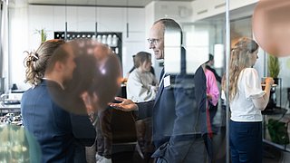 Foto, mehrere Personen in einem modernen Büro stehen und unterhalten sich in kleinen Gruppen.