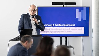 Foto, Mann mit Mikrofon vor Bildschirm mit Präsentationstext, im Vordergrund unscharfe Personen.