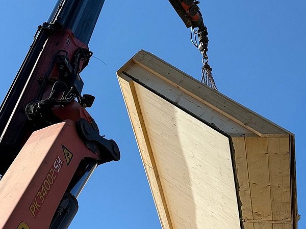 Foto, Kran hebt große rechteckige Holzplatte mit Metallrahmen vor blauem Himmel