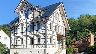 Foto, Fachwerkhaus mit weißen Wänden und braunen Holzbalken, Solarpaneele auf dem Dach, Treppe zum Eingang, Autos und Bäume im Hintergrund