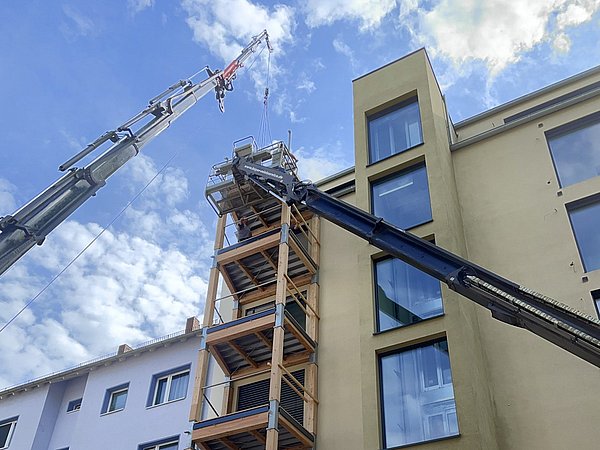 Foto, an einem Mehrfamilienhaus werden von zwei Kränen Balkone aus Holz angebracht
