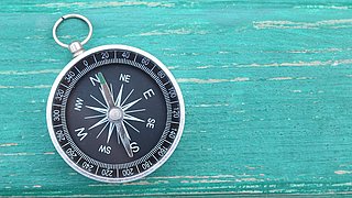 Foto, analoger Taschen-Kompass, der auf einer grün lackierten Holzoberfläche liegt.