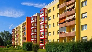 Foto, mehrere gelbe Wohngebäude mit roten Treppenhäusern und Balkonen, davor grüne Büsche und Rasenfläche unter blauem Himmel.