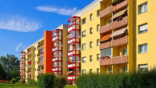 Foto, mehrere gelbe Wohngebäude mit roten Treppenhäusern und Balkonen, davor grüne Büsche und Rasenfläche unter blauem Himmel.