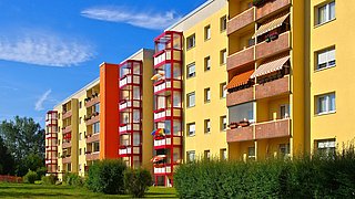 Foto, mehrere gelbe Wohngebäude mit roten Treppenhäusern und Balkonen, davor grüne Büsche und Rasenfläche unter blauem Himmel.