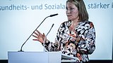 Foto, Person mit schulterlangem Haar und gemusterter Bluse spricht an einem Rednerpult mit Mikrofon vor einer Präsentationsfolie mit Text
