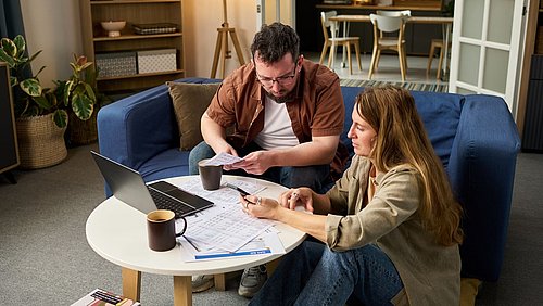 Foto, zwei Personen sitzen auf einem blauen Sofa und bearbeiten gemeinsam Dokumente an einem runden Tisch mit Laptop, Kaffeetassen und Papieren in einem wohnlich eingerichteten Raum.