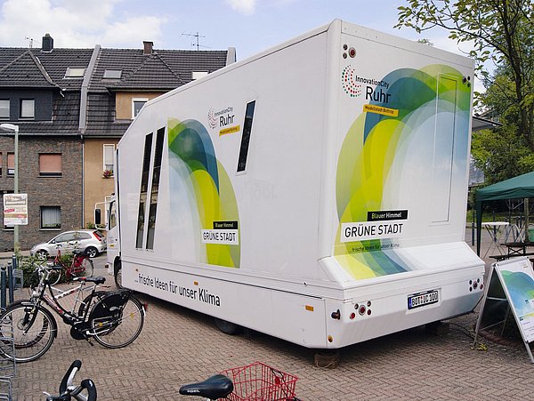 Foto, Weißes Infomobil mit Aufschrift 'InnovationCity Ruhr' und 'Grüne Stadt' steht auf gepflastertem Platz vor Wohnhäusern