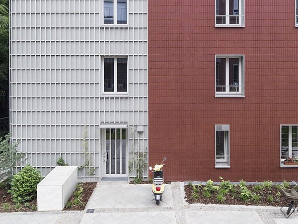 Zwei Wohngebäude mit unterschiedlichen Fassaden: links grau mit vertikalen Paneelen, rechts rot mit quadratischen Kacheln. Vor dem Gebäude stehen ein Fahrrad und ein elektrischer Roller.