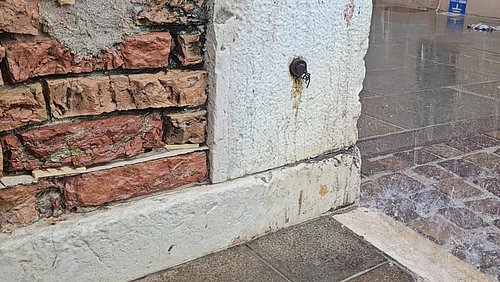 Foto, freigelegtes Mauerwerk aus roten Ziegelsteinen neben einem verputzten Sockel mit Wasser, das auf den Boden spritzt.