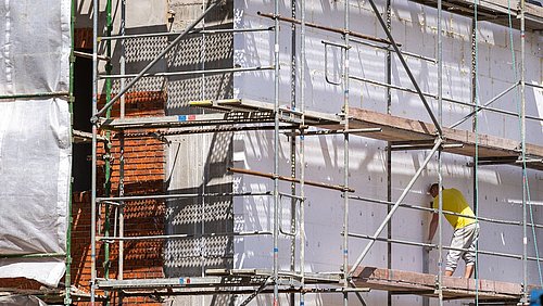 Foto, Baugerüst vor einem Haus mit teilweise sichtbarer roter Backsteinwand und einem Mann in gelbem T-Shirt, der an der weißen Fassade arbeitet.