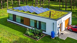 Foto, flaches modernes Haus mit begrüntem Dach und Solarpaneelen, umgeben von Rasen und Wald, mit kleinem Gemüsegarten und rotem Auto neben Garage