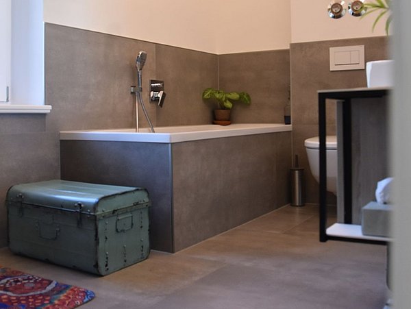 Foto, Badezimmer mit grauen Fließen und weißen Wänden. Im Zentrum befindet sich eine Badewanne, rechts ein Waschtisch, links eine antike Truhe.