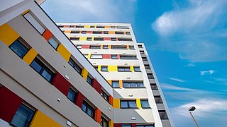 Foto, Blick nach oben auf ein mehrstöckiges Wohngebäude mit farbigen Fassadenelementen in Gelb, Rot und Orange unter blauem Himmel mit Wolken.