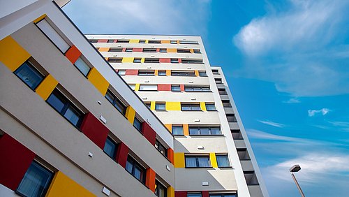 Foto, Blick nach oben auf ein mehrstöckiges Wohngebäude mit farbigen Fassadenelementen in Gelb, Rot und Orange unter blauem Himmel mit Wolken.