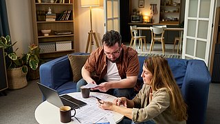 Foto, Ein Mann und eine Frau sitzen gemeinsam an einem runden weißen Couchtisch in einem Wohnzimmer. Beide schauen konzentriert auf Dokumente und einen Laptop, der auf dem Tisch steht. Die Frau hält einen Stift und zeigt auf ein Blatt Papier, während der Mann weitere Unterlagen in der Hand hält. Im Hintergrund sind ein Bücherregal, eine Stehlampe, Pflanzen und eine offene Tür zu einer Küche sichtbar.