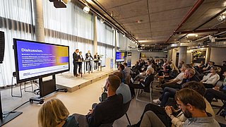 Foto, eine Podiumsdiskussion in einem modernen Veranstaltungsort mit engagierten Teilnehmern, bei der die Redner vor großen Bildschirmen sitzen, auf denen das Thema dargestellt wird.