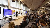 Foto, eine Podiumsdiskussion in einem modernen Veranstaltungsort mit engagierten Teilnehmern, bei der die Redner vor großen Bildschirmen sitzen, auf denen das Thema dargestellt wird.