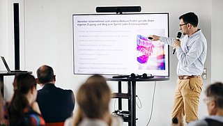 Foto, Mann präsentiert vor Publikum mit Mikrofon und zeigt auf Bildschirm mit Text und farbigem Bild bei einer Veranstaltung.