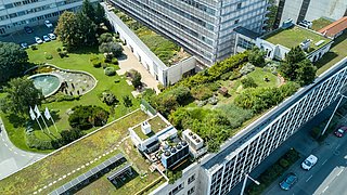Foto, Luftaufnahme eines modernen Gebäudes mit grünen Dächern, Gärten und einem Teich, umgeben von Bäumen und geparkten Autos.
