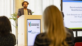 Foto, ein Redner hält einen Vortrag auf einer Konferenz, während hinter ihm ein großer Bildschirm mit Text über nachhaltige Baupraktiken zu sehen ist.