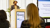 Foto, ein Redner hält einen Vortrag auf einer Konferenz, während hinter ihm ein großer Bildschirm mit Text über nachhaltige Baupraktiken zu sehen ist.