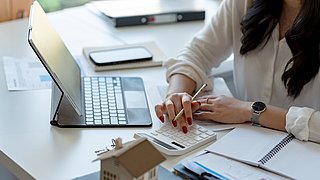 Foto, Frau tippt auf Taschenrechner neben Tablet und Unterlagen auf Schreibtisch.