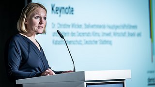 Foto, Frau mit blauer Bluse steht an einem weißen Rednerpult mit Mikrofon, im Hintergrund unscharfe Projektion.