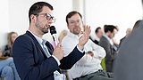 Foto, Mann mit Brille hält Mikrofon und spricht in einem Publikum mit mehreren Personen im Hintergrund.